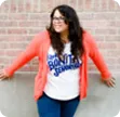 Smiling woman with glasses, wearing a white graphic t-shirt and red cardigan, standing against a brick wall.